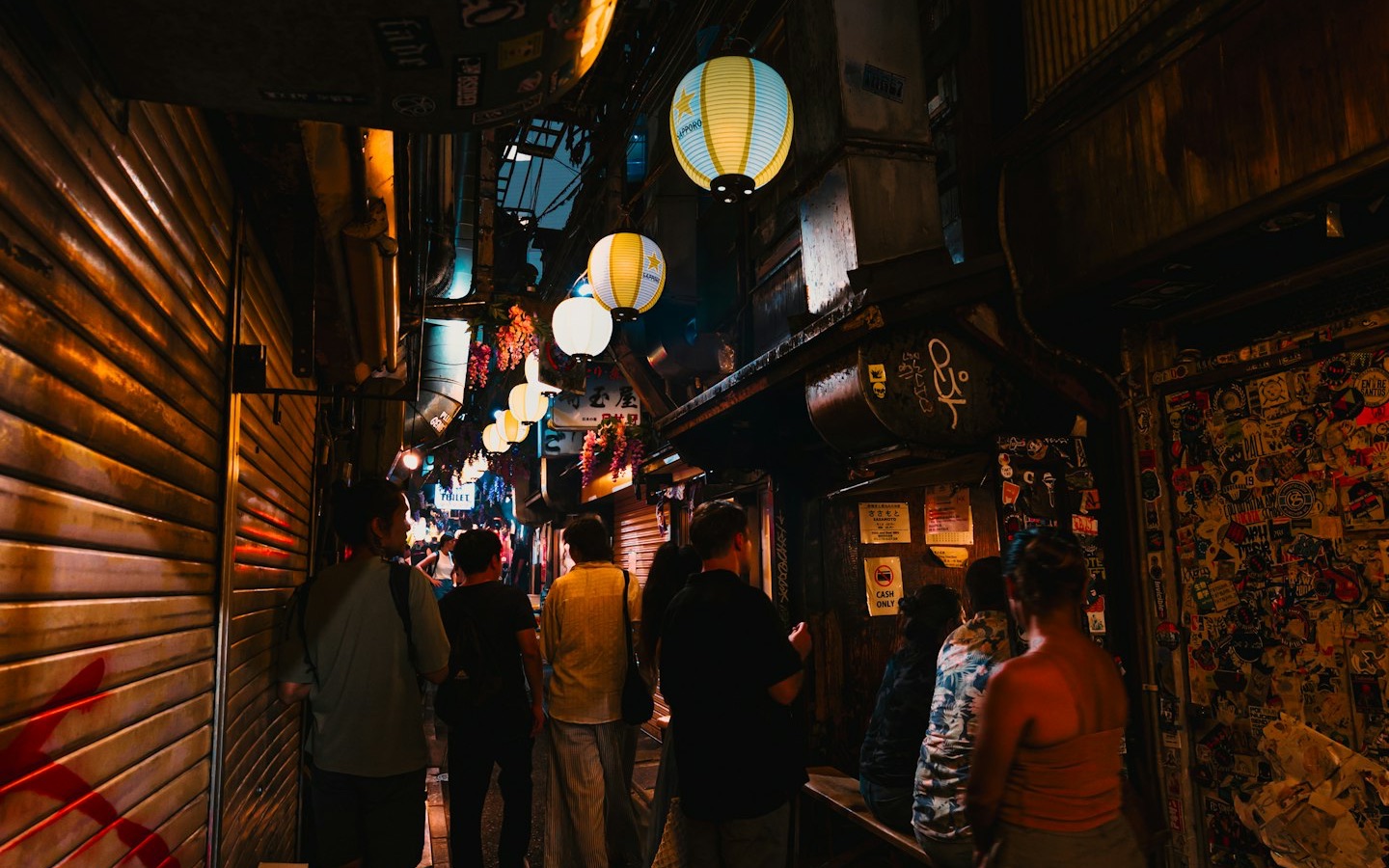A narrow Japanese alley at night lined with small shops and glowing paper lanterns