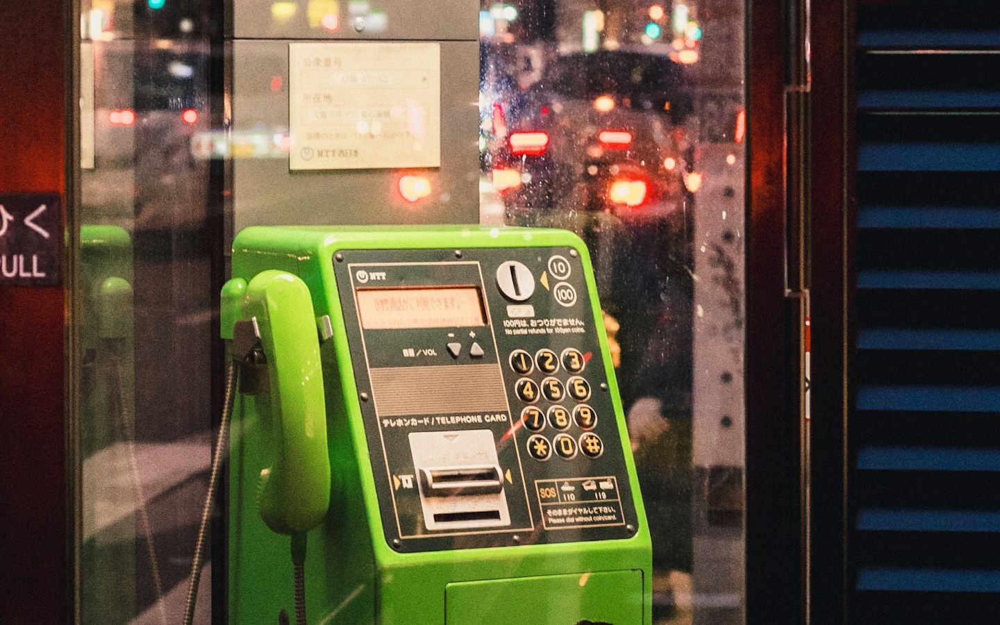 A bright green NTT public payphone in a glass phone booth on a Tokyo street at night